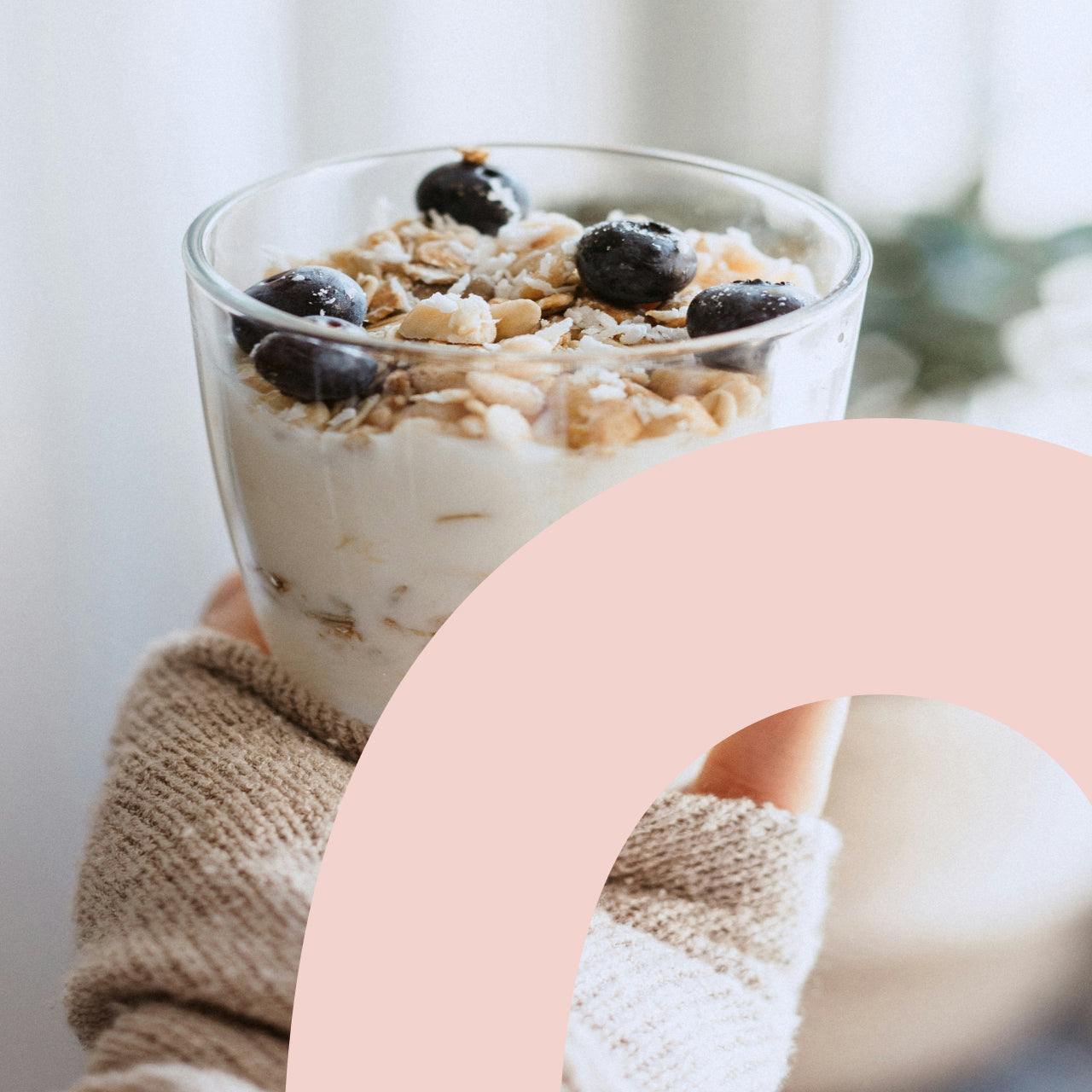 Glass of yogurt parfait with granola and fresh blueberries held by hands in a beige knit sweater, partially framed by a pink curved Bodywealth logo element, with a soft blurred background.